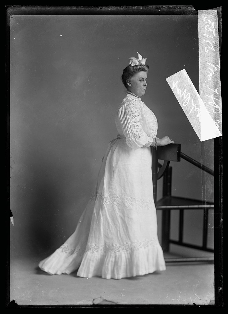 Full-length portrait of a woman in a white gown with a train, standing beside a chair, photographed by C. M. Bell, identified as Mrs. H. P. Marston.