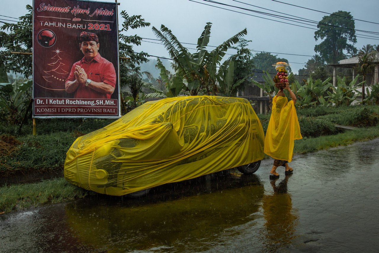 A woman in a yellow raincoat carries offerings past a yellow car cover and a political poster in the rain, Bali