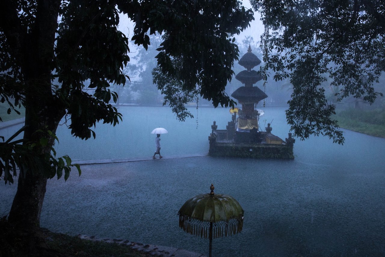 A lone figure with a white umbrella walks through a flooded temple courtyard in heavy rain, Bali