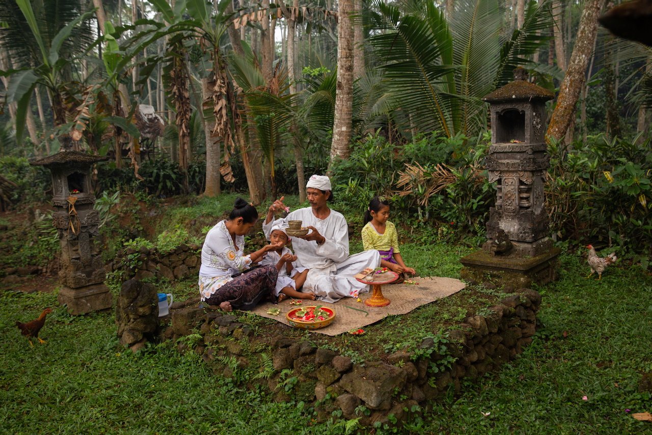 A family conducts a ceremony on a stone platform surrounded by jungle shrines, Bali