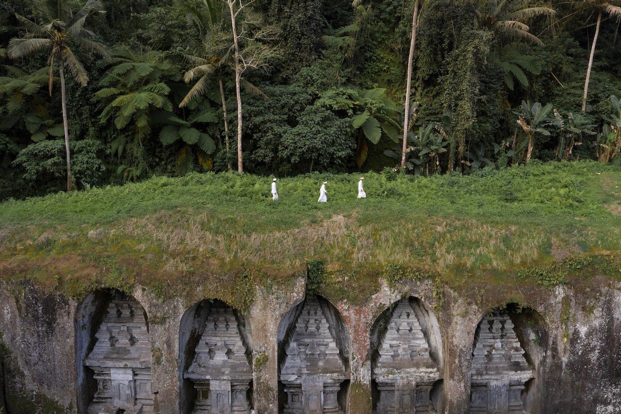 Three small white figures walk above a row of carved stone niches in the jungle, Bali