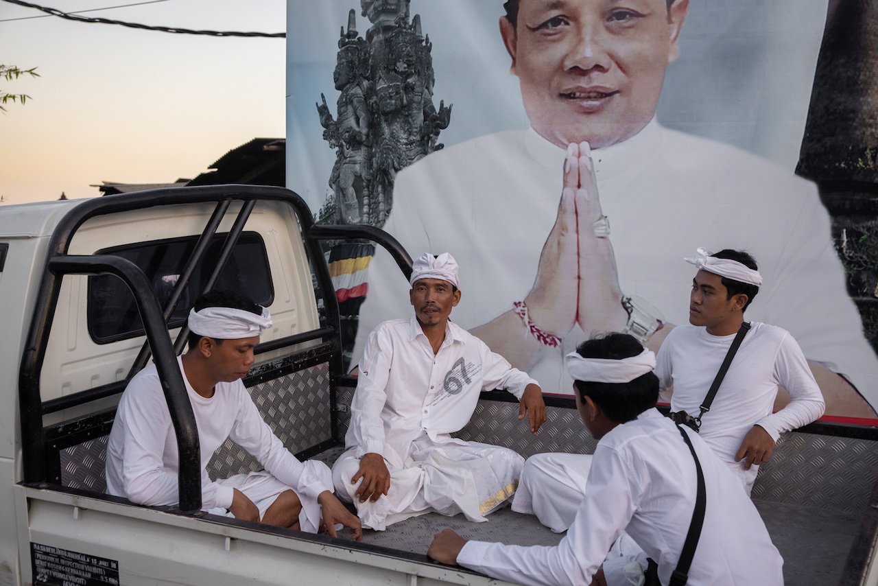 Men in white temple dress sit in the back of a truck in front of a large political campaign poster, Bali