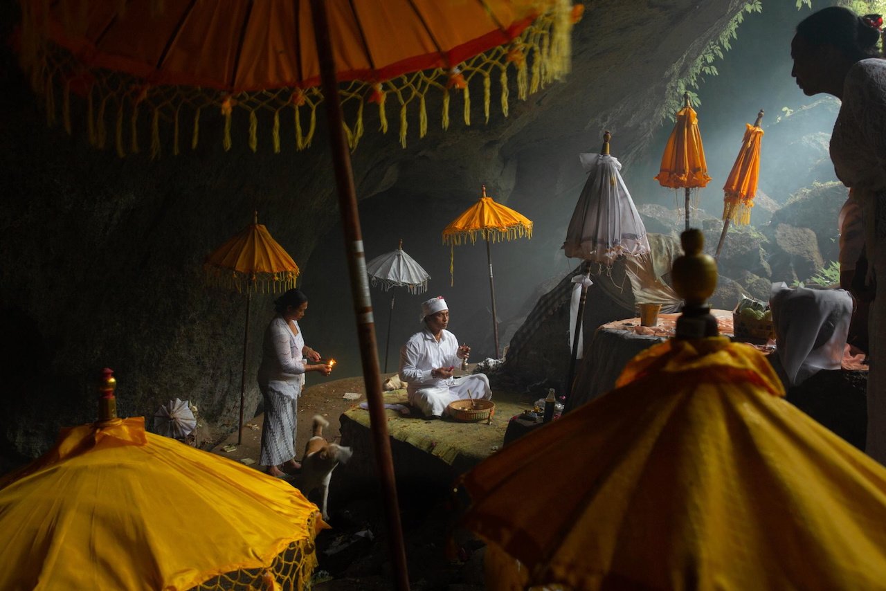 A priest prepares offerings beneath ceremonial umbrellas inside a cave temple, Munduk, Bali