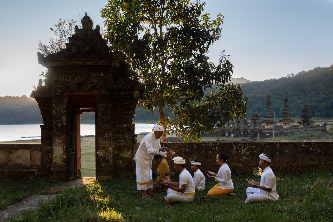 A priest administers holy water to worshippers seated on grass beside a lake temple, Bali