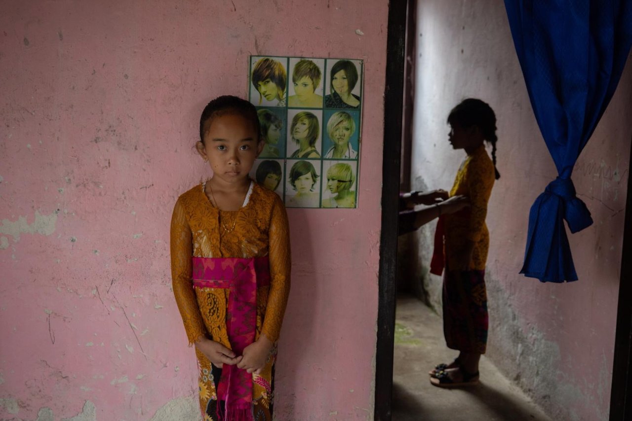 A young girl in traditional Balinese dress stands before a Western hairstyle poster on a pink wall, Singaraja, Bali