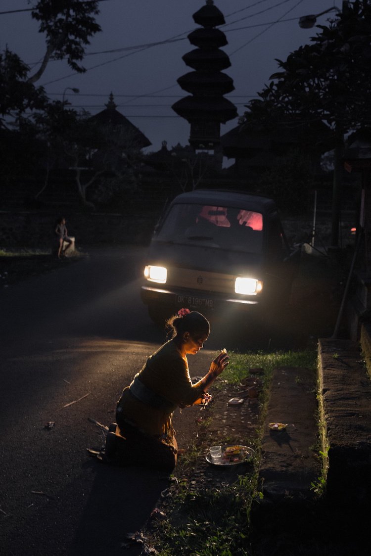 A woman makes an offering at the roadside at night, lit by oncoming headlights, a temple tower rising in the darkness behind her, Bali